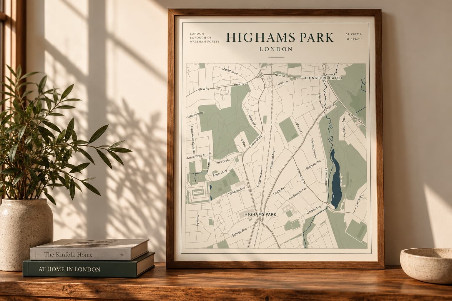 A framed A4 map print of Highams Park, London, leaning on an oak shelf beside a plant, books and a ceramic bowl in warm window light